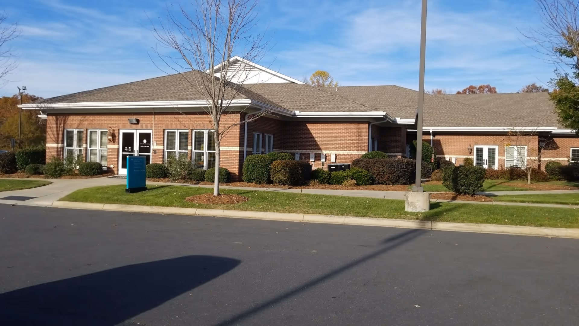 Single-story brick hospice building with windows, shrubs, and a paved entrance under a blue sky.