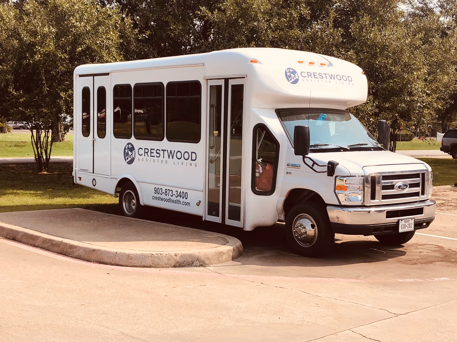 A white Crestwood assisted living bus parked beside trees on a curb.