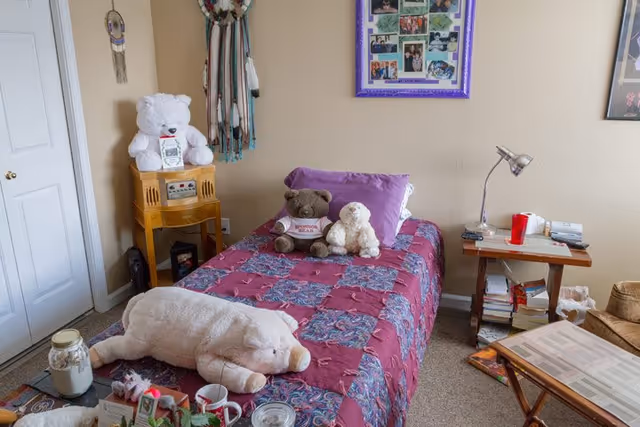 A cozy bedroom with a single bed covered in a red and blue patterned quilt. On the bed are three stuffed animals: a large white pig, a brown teddy bear wearing a shirt, and a white lamb. To the left of the bed is a small wooden cabinet with a white teddy bear on top and a dreamcatcher hanging on the wall above it. On the right side of the bed is a wooden side table with a silver desk lamp, a red cup, and some books. The walls are beige with framed photos and artwork hanging. There is a closet door on the left side of the room.