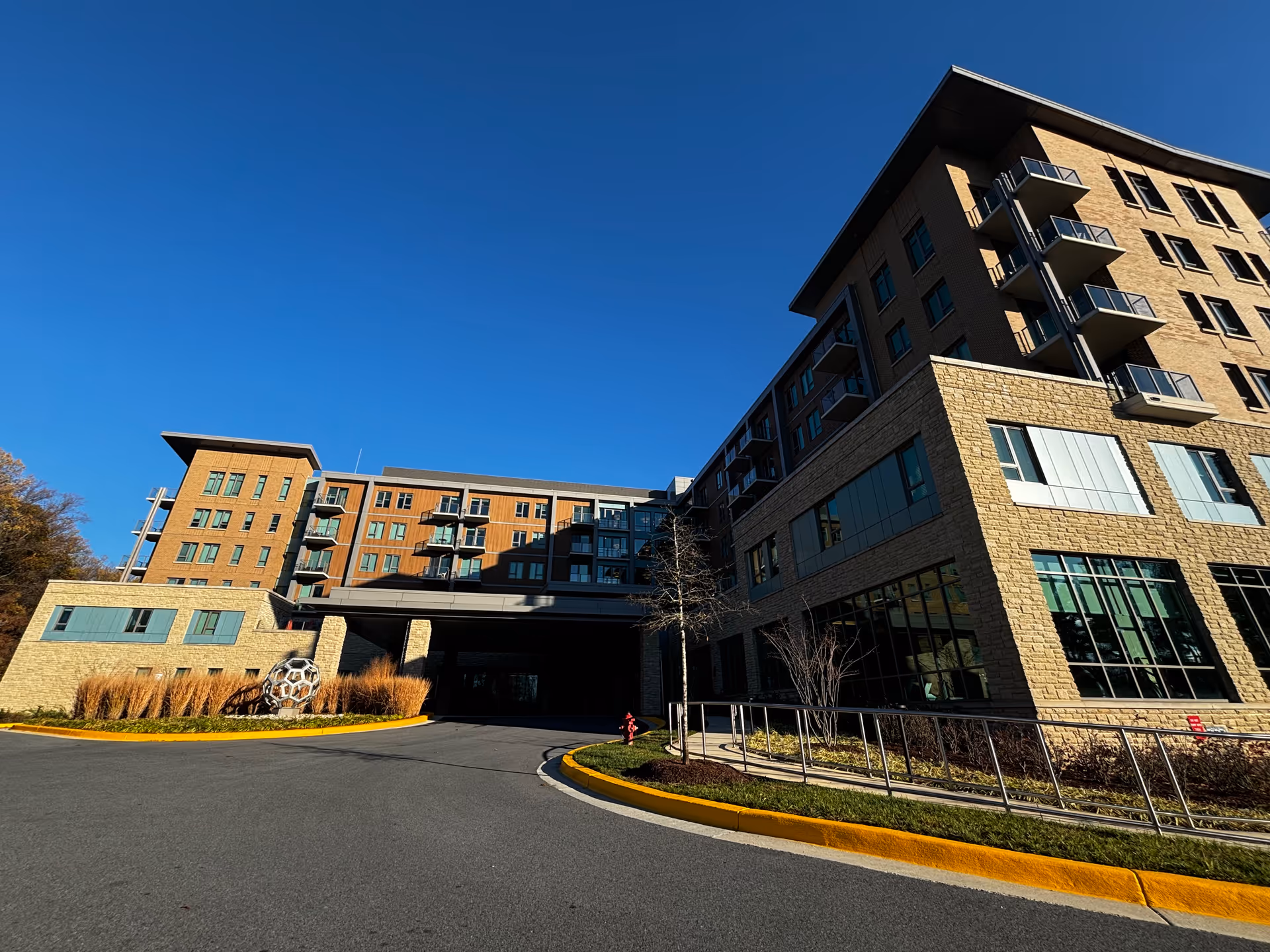 Front exterior of a multi-story senior living building with balconies, an entrance canopy and a curved driveway under a clear blue sky.