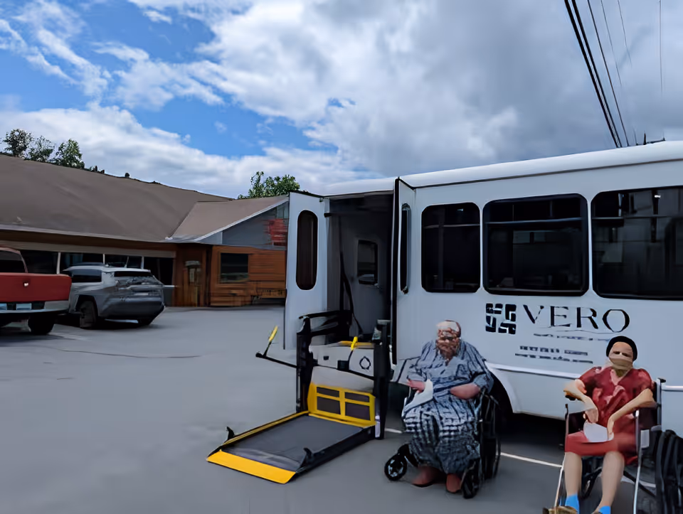 White Vero shuttle with its wheelchair lift deployed in the parking lot in front of a single-story senior living building, two people in wheelchairs nearby.