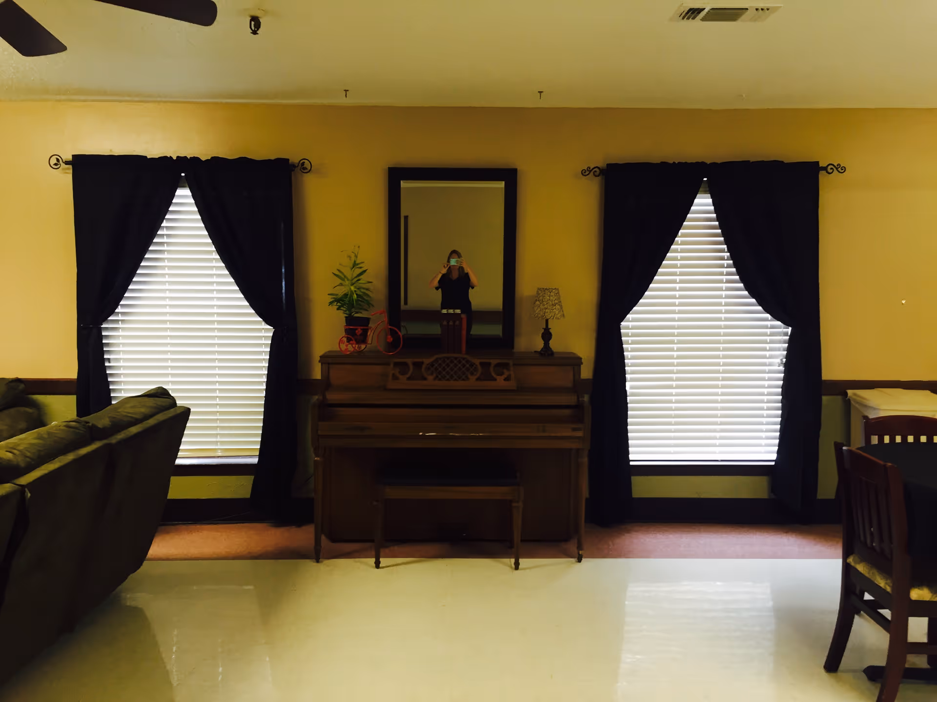 Interior view of a living room area in Kaufman Healthcare Center featuring a wooden piano with a bench, a mirror above it, two windows with black curtains and white blinds, a small table lamp, a potted plant, a green sofa on the left, and a dining table with chairs on the right.