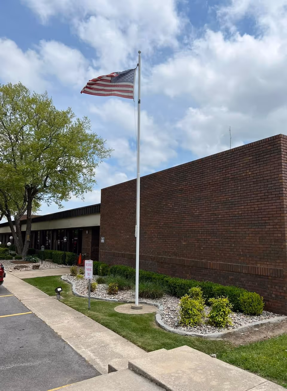 Exterior view of Ballard Nursing Center showing a brick building with a flagpole flying an American flag, a tree, landscaped bushes, and a parking area with a reserved parking sign.