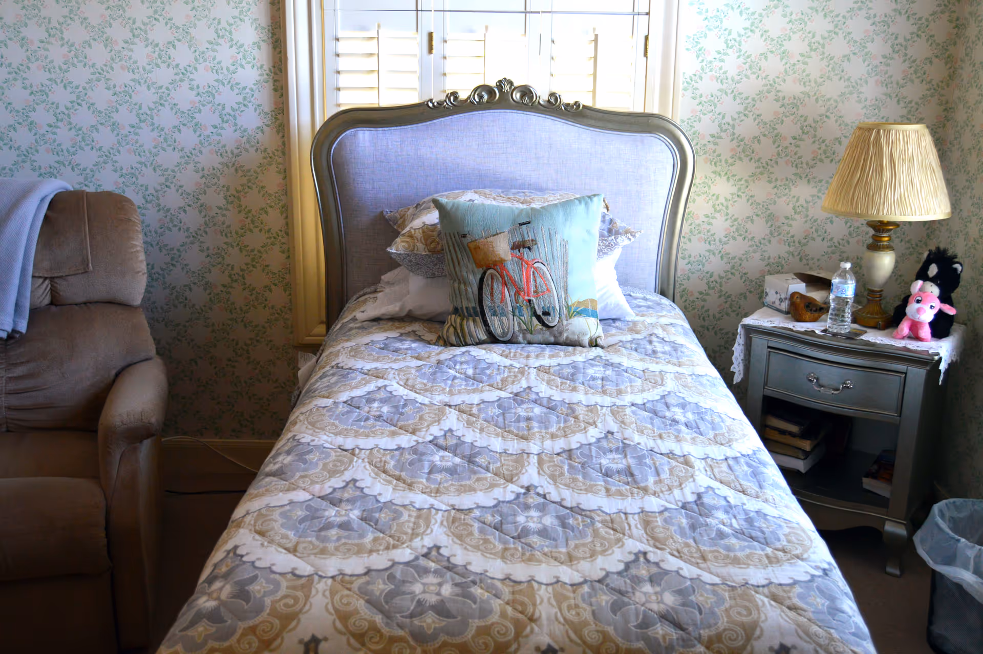 A bedroom with a tufted headboard bed, nightstand, lamp, and floral wallpaper.