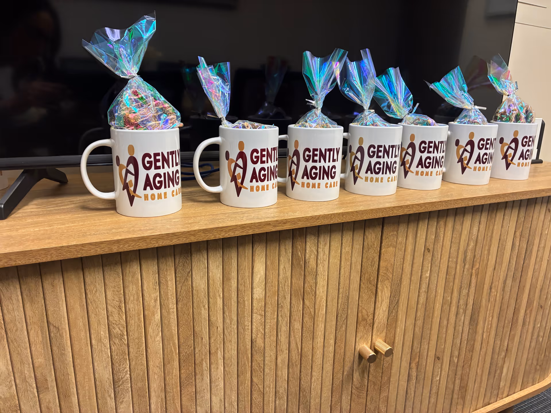 Row of mugs branded 'Gently Aging Home Care' with wrapped candies on a wooden reception counter.