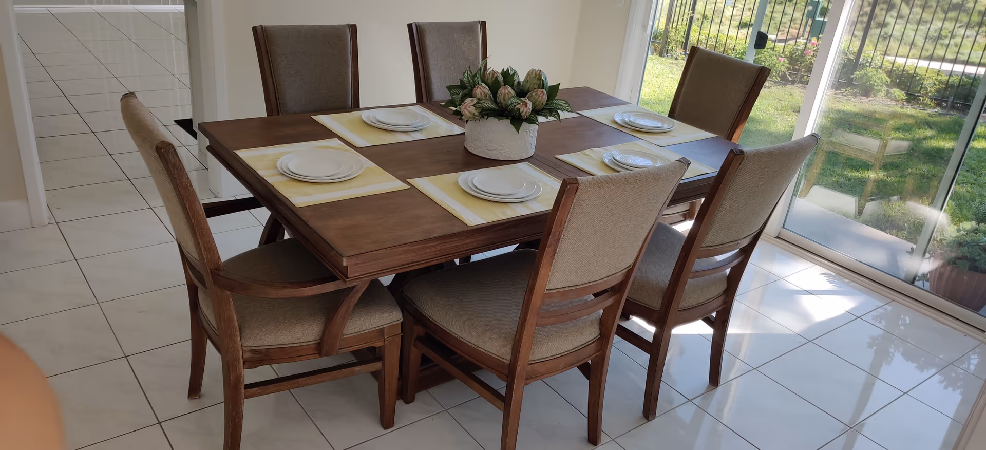 A wooden dining table with six chairs set with plates in a bright, tiled dining area.