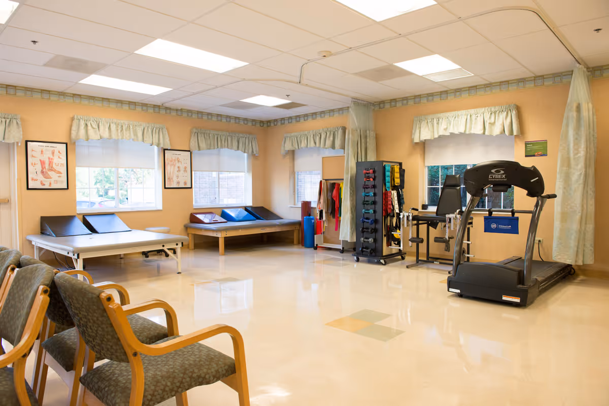A rehabilitation gym in a pale-walled facility featuring treadmills, weights, tables, and waiting chairs.
