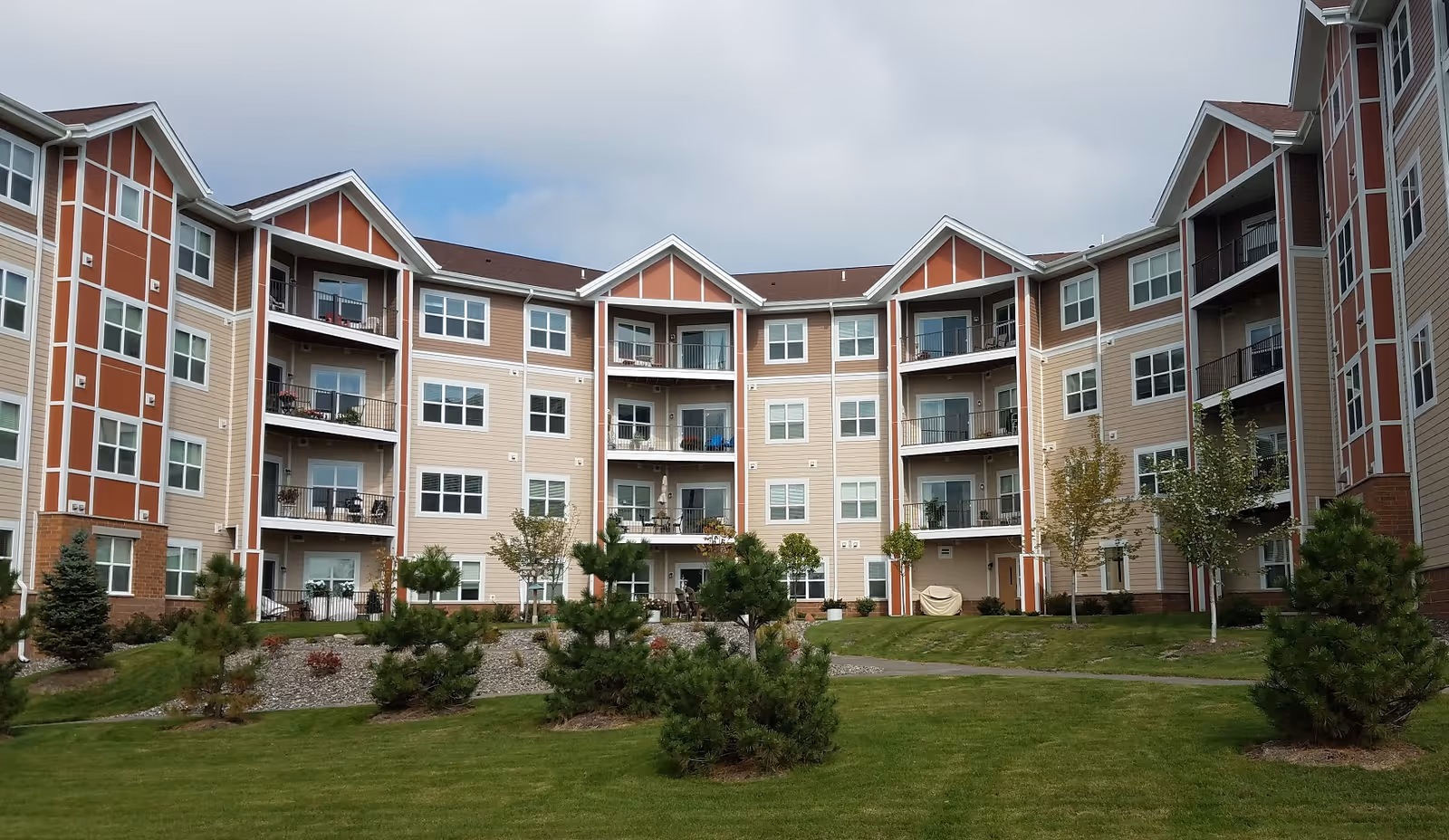 Exterior view of a multi-story senior living community building with balconies, windows, and a well-maintained lawn with small trees and shrubs in front under a partly cloudy sky.