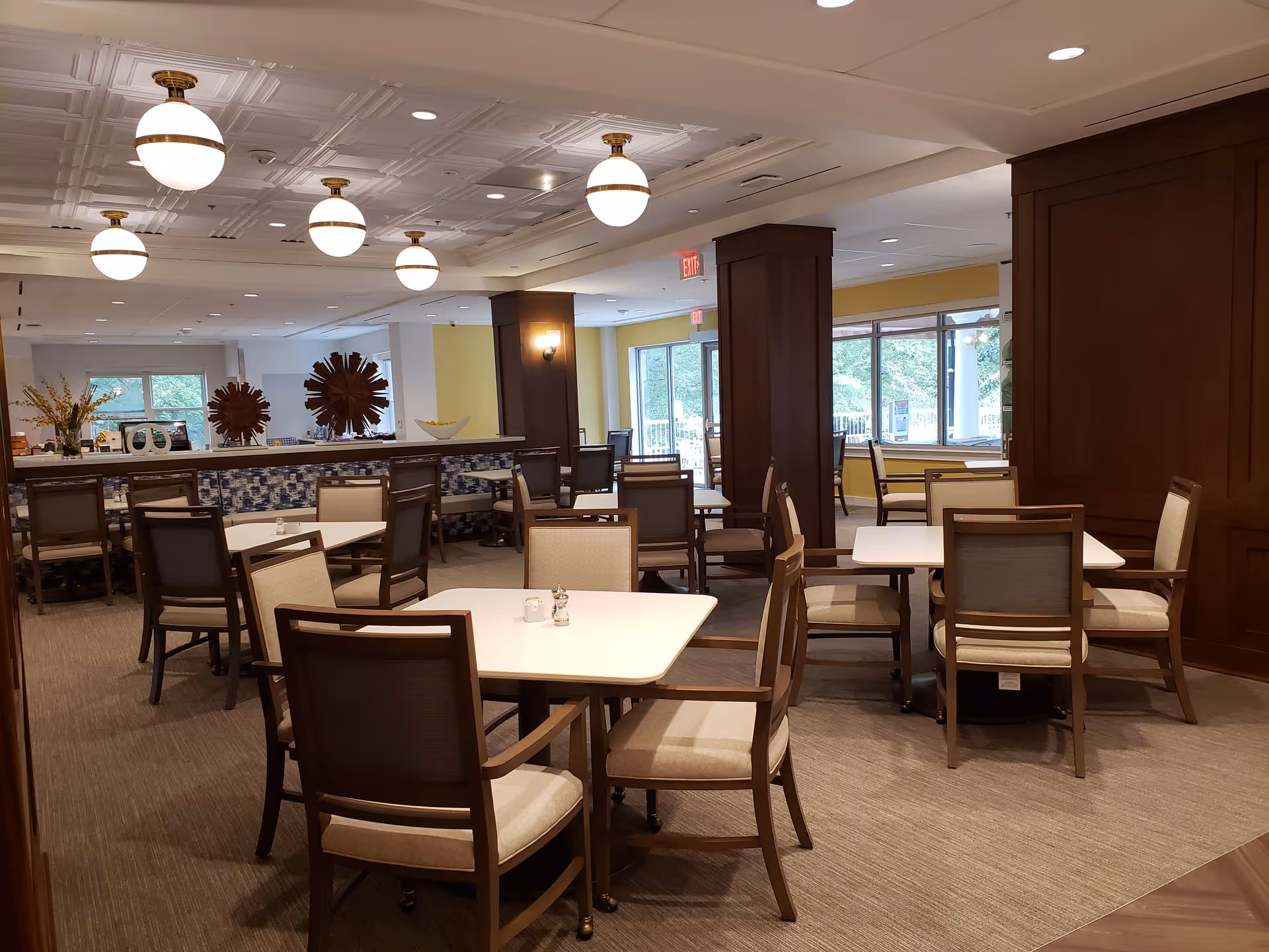 A well-lit dining room with multiple square tables and cushioned chairs arranged neatly. The room features large windows allowing natural light to enter, decorative wooden columns, and ceiling lights with round fixtures. There are decorative items on a counter in the background and a view of greenery outside the windows.