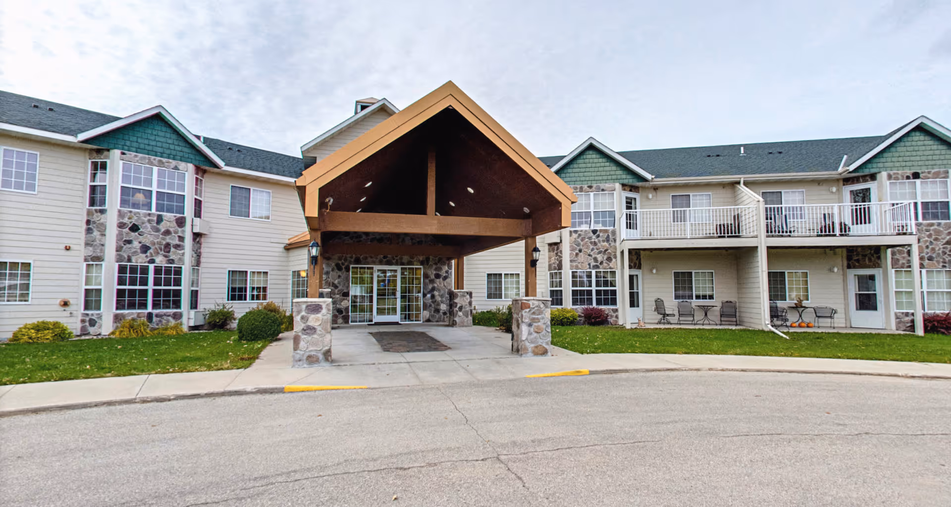 Front exterior view of Tamarac Senior Living facility showing a two-story building with stone and siding facade, a covered entrance with wooden beams, multiple windows, and outdoor seating areas on the ground and upper floors.