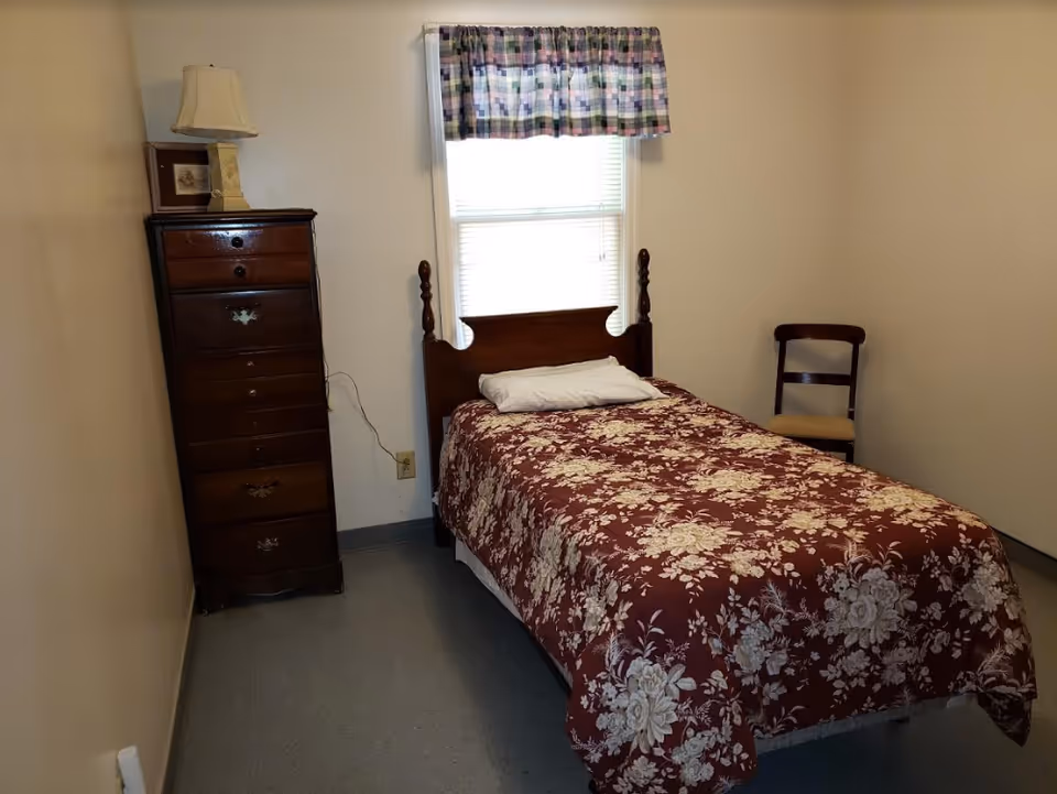 Small bedroom with single bed, floral quilt, dresser, window, and chair.