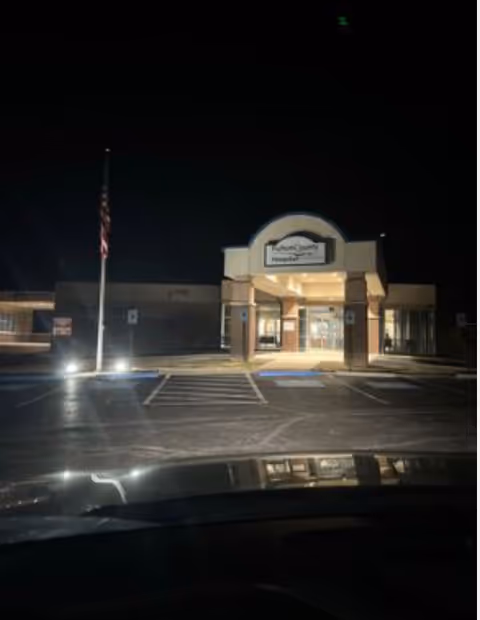 Night exterior of a hospital entrance building with parking lot and flagpole.