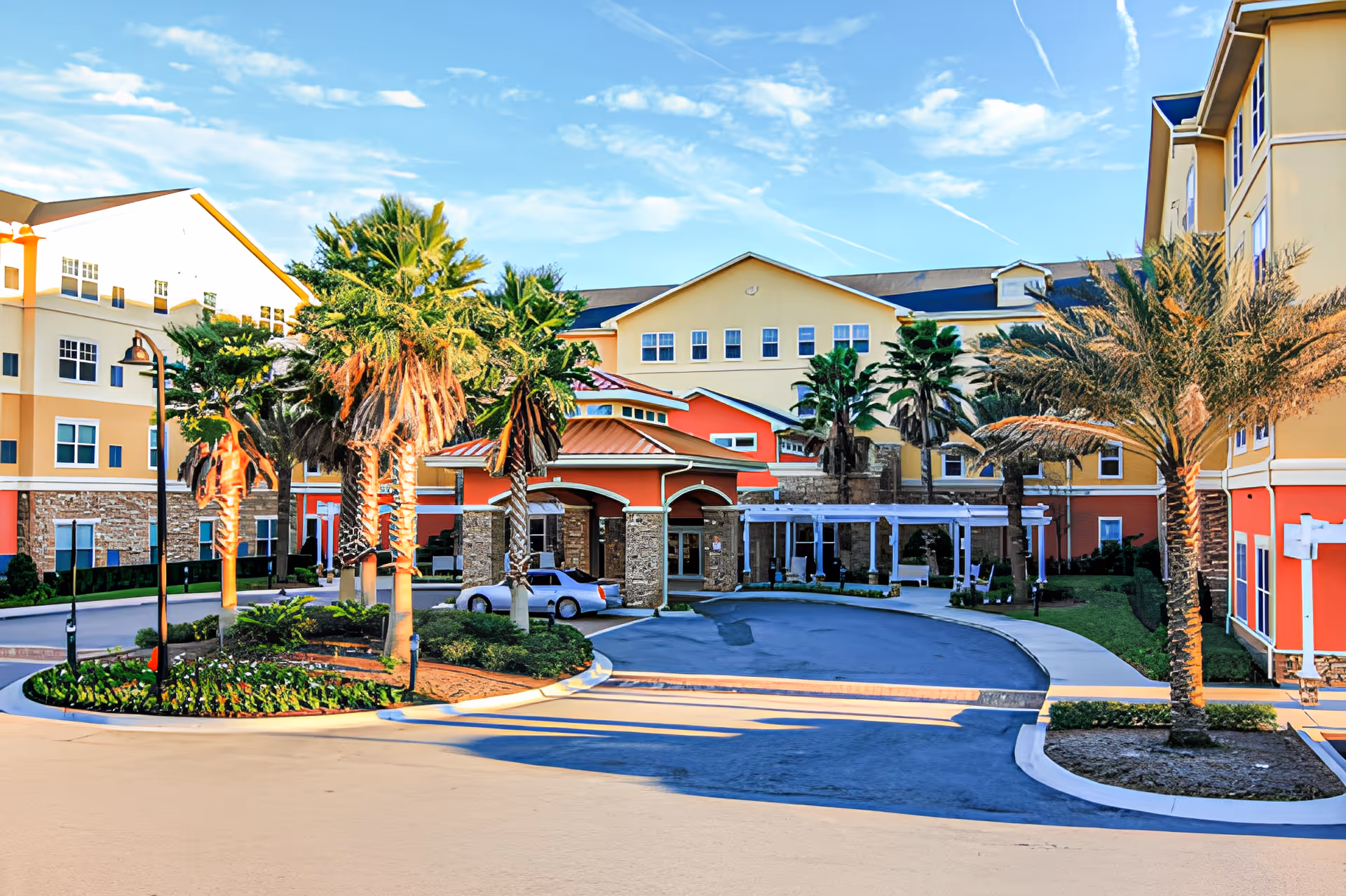 Exterior view of Camellia At Deerwood senior living facility featuring a circular driveway with palm trees and landscaped greenery, multi-story buildings with yellow and orange walls, and a clear blue sky with scattered clouds.