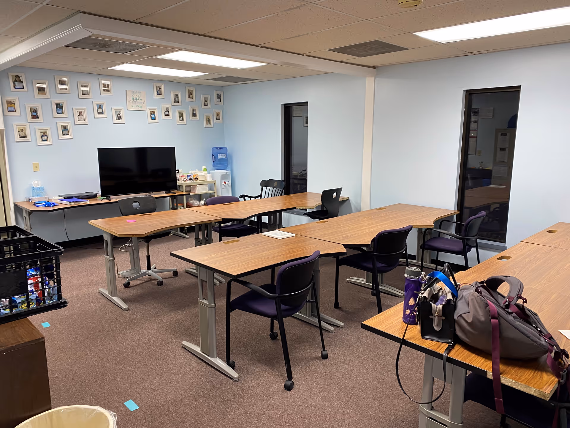 A light blue-walled activity room with rectangular tables, chairs, and a TV.
