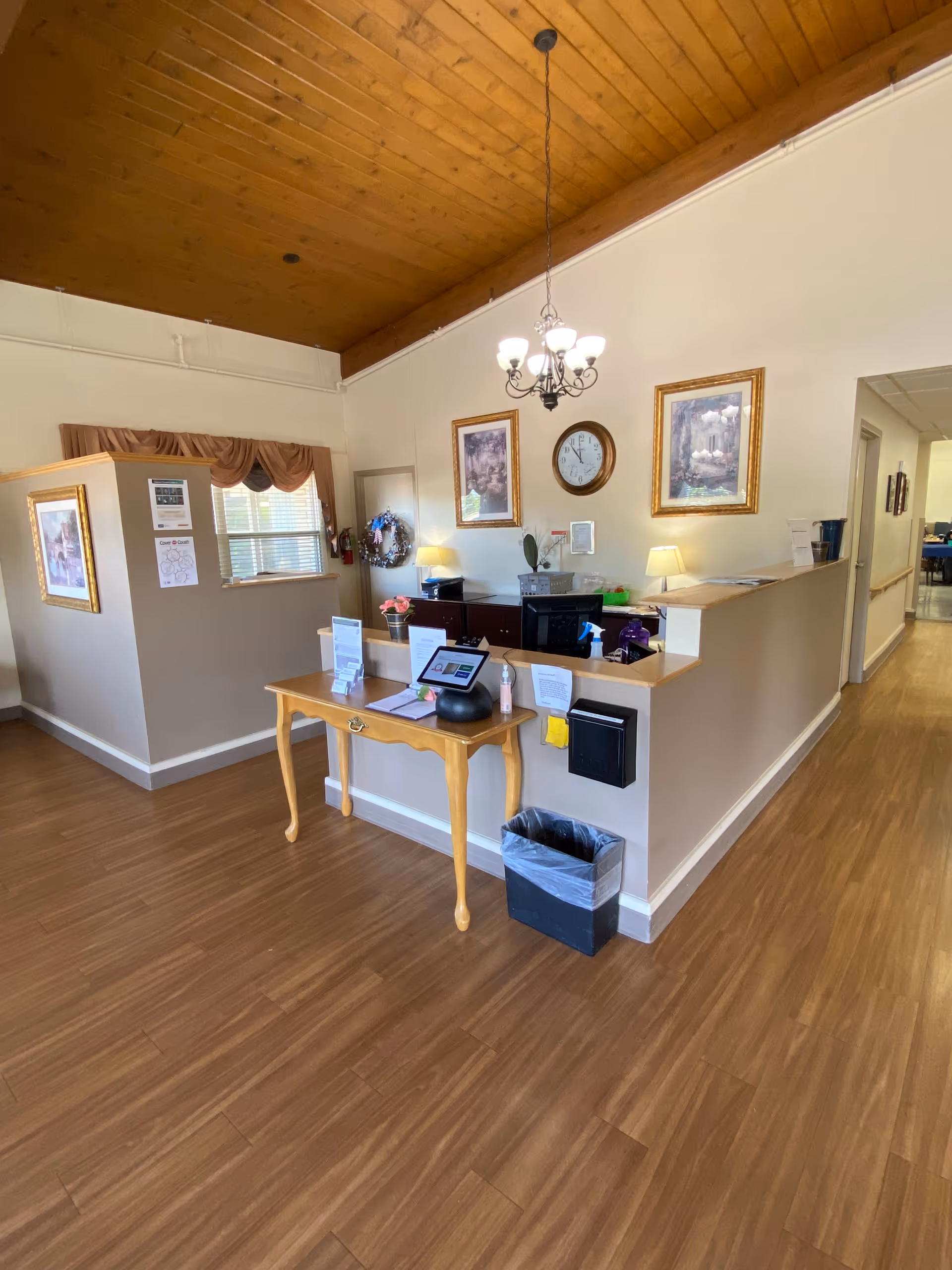Interior view of a reception or front desk area in a senior living facility with wooden flooring and a wooden ceiling. A small wooden table with informational materials and a tablet is placed in front of a half wall counter. Behind the counter are two lamps, framed pictures, a clock, and various office supplies. A hallway extends to the right.