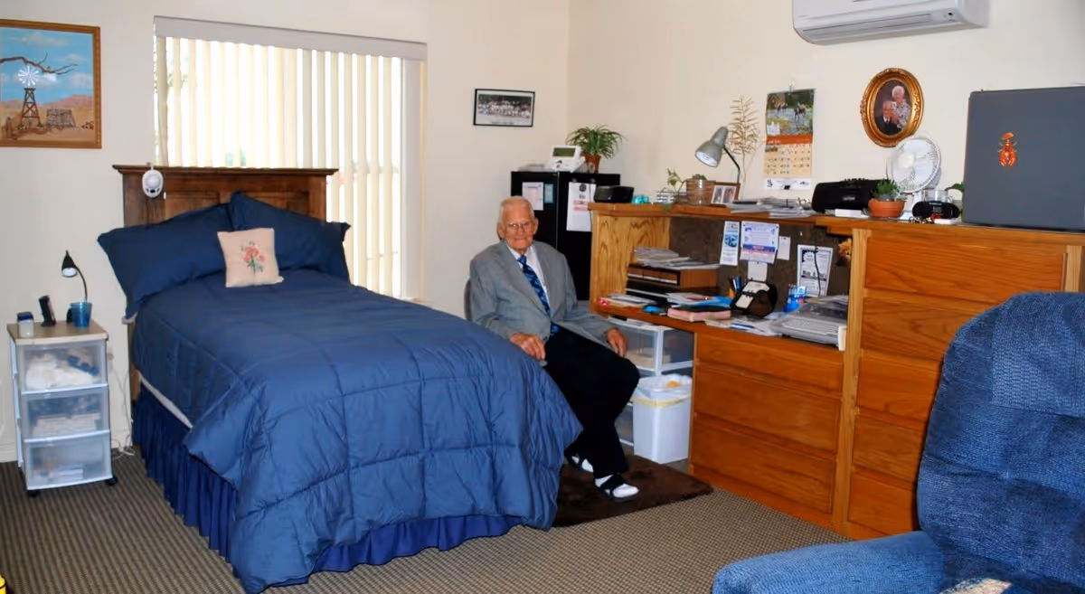 A senior man wearing a gray suit jacket and blue tie is sitting on a chair in a bedroom. The room features a bed with a blue comforter and a floral pillow, a wooden headboard, a small white plastic drawer unit beside the bed, a wooden dresser with various items on top including a lamp, calendar, fan, and framed photos. There is a blue upholstered chair in the foreground and vertical blinds covering a window behind the bed.