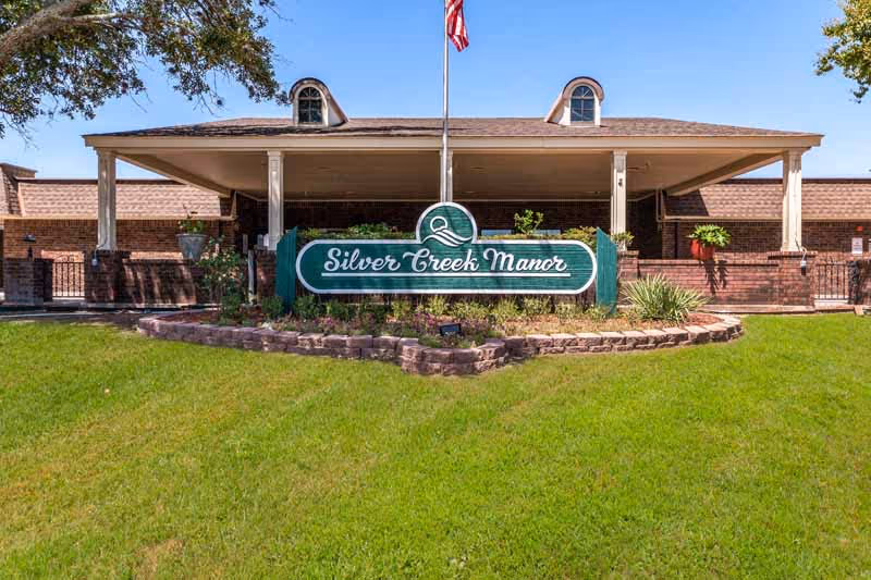 Front entrance of Silver Creek Manor showing a green facility sign on a landscaped lawn beneath a covered portico and an American flag.