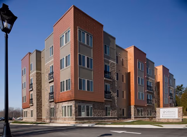 Exterior view of a modern multi-story senior living facility building with a mix of stone and reddish-brown paneling under a clear blue sky. A street lamp is visible on the left, and a sign for The Greens at Liberty Road is on the right near the entrance driveway.