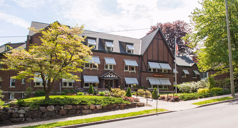 Brick multi-story home with gabled roof, window awnings, landscaped front yard and an American flag.