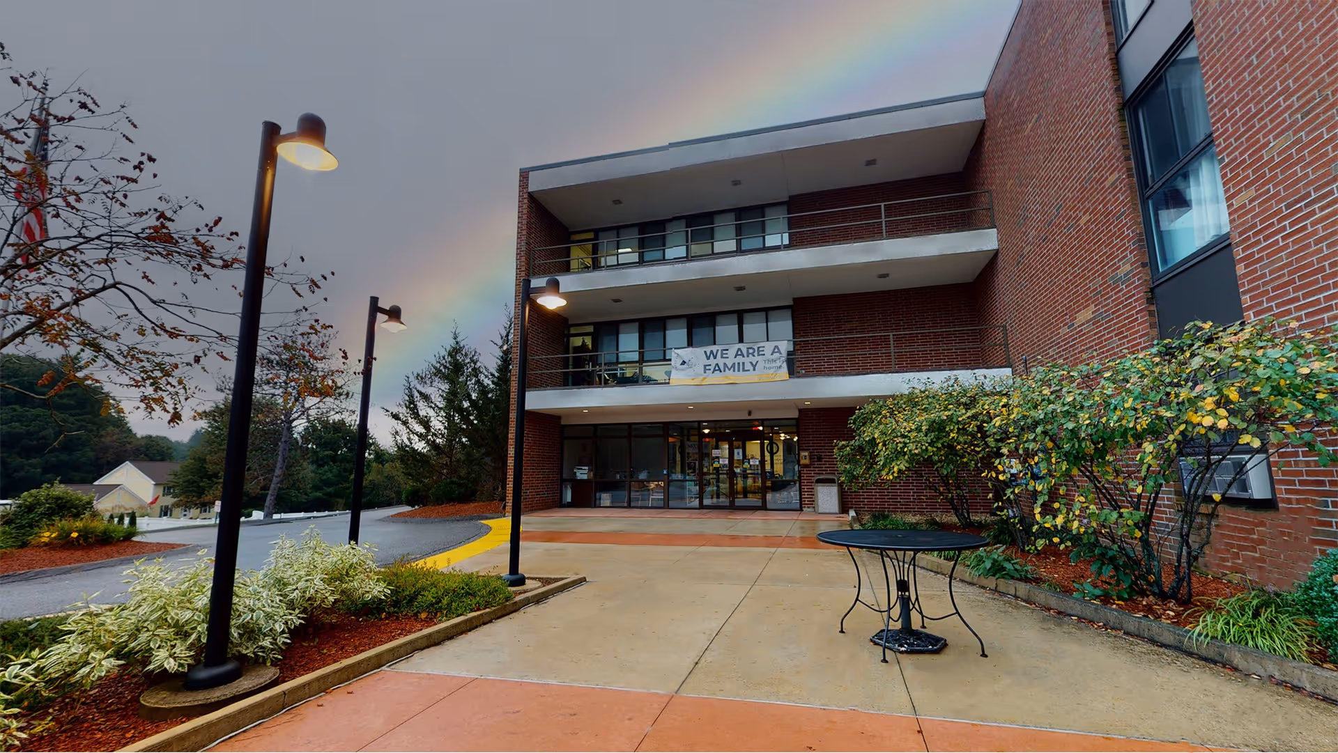 Front entrance of a three-story brick senior living building with a banner reading "WE ARE A FAMILY", outdoor lamps, a small patio table, and a rainbow in the sky.
