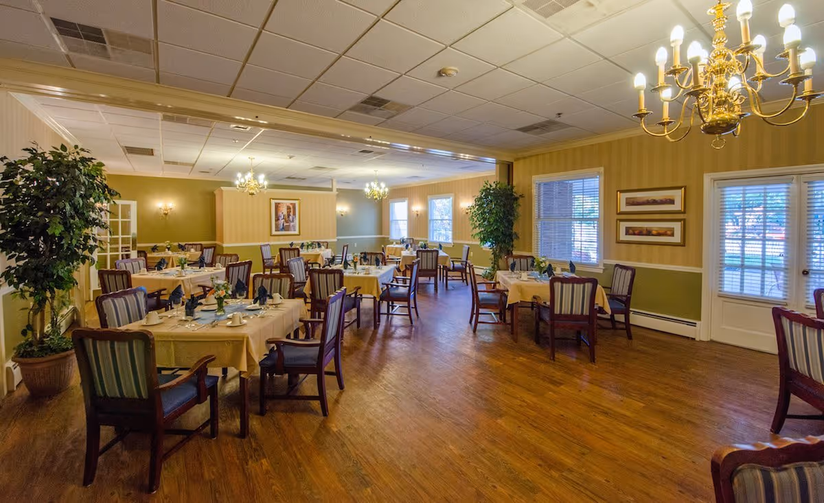 Senior living facility dining room with several tables set for meals, chairs, plants, and chandeliers.