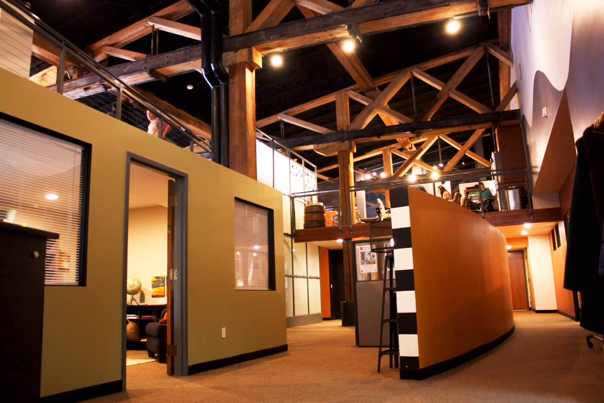 Open, modern interior with exposed wooden beams, orange curved wall, upper mezzanine, and doorway.