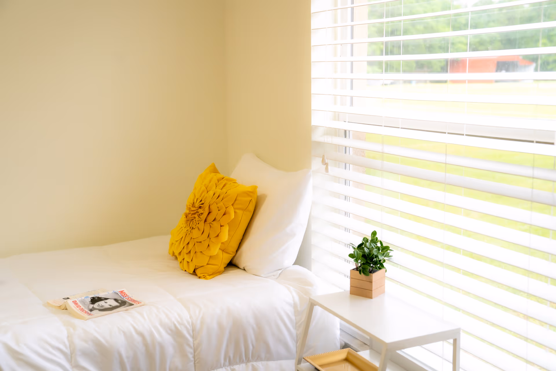 Bright bedroom with white bedding, a yellow pillow, a small plant on a table by blinds.