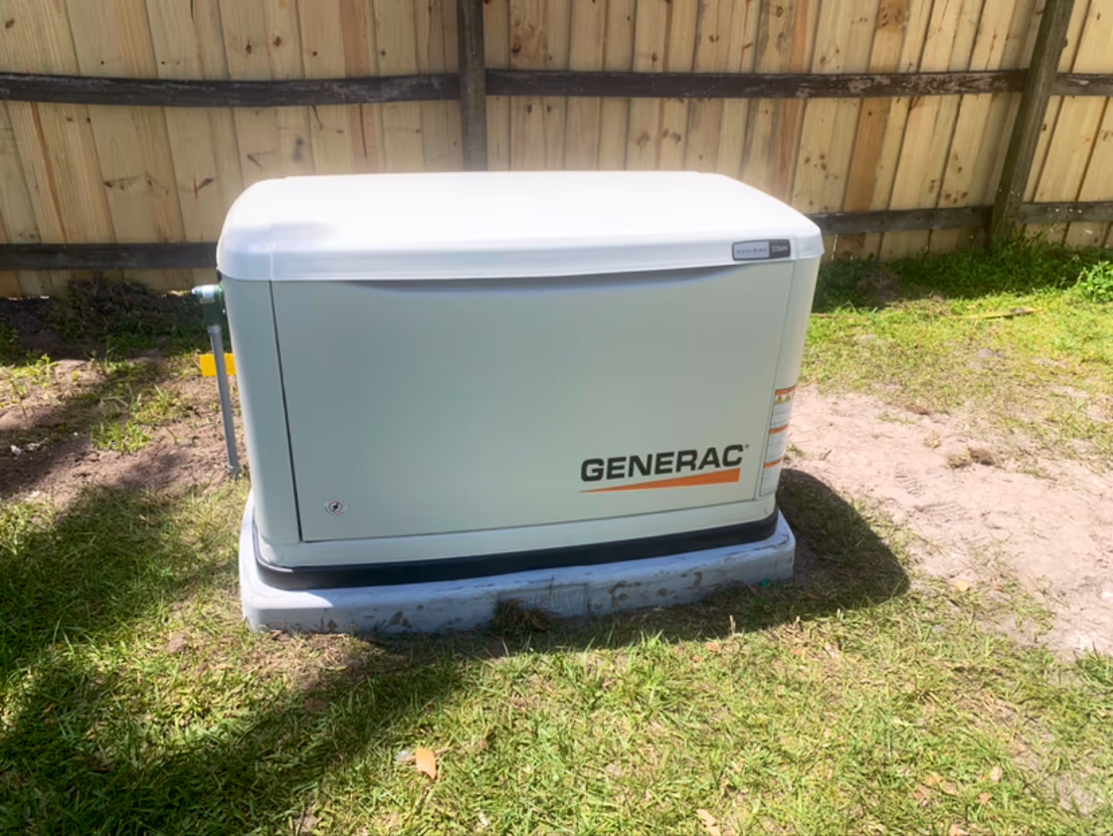 A Generac standby generator sits on a concrete pad beside a wooden fence.
