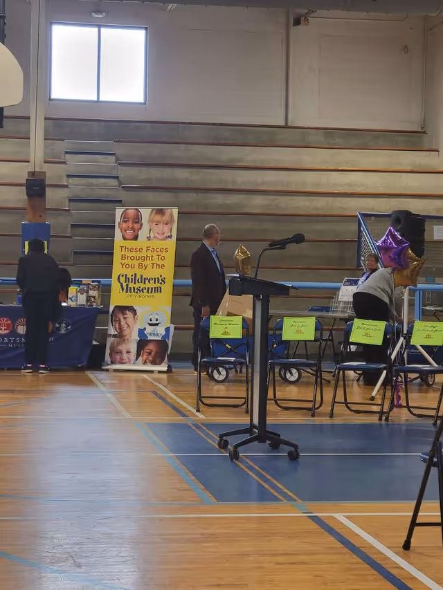 Indoor gym with a podium, banners, tables, and people preparing for an event