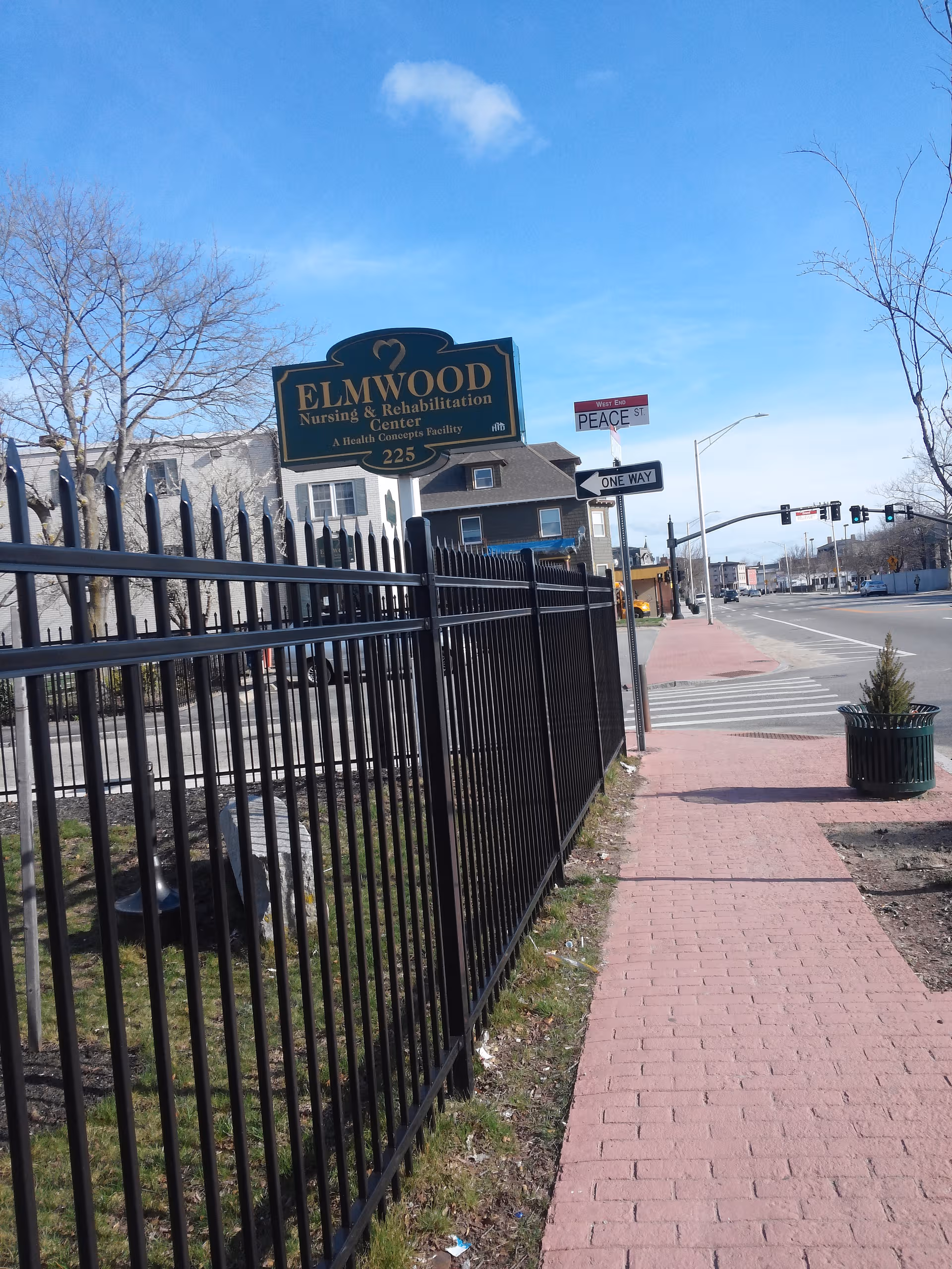 Elmwood Nursing & Rehabilitation Center sign beside a black fence on a city street.