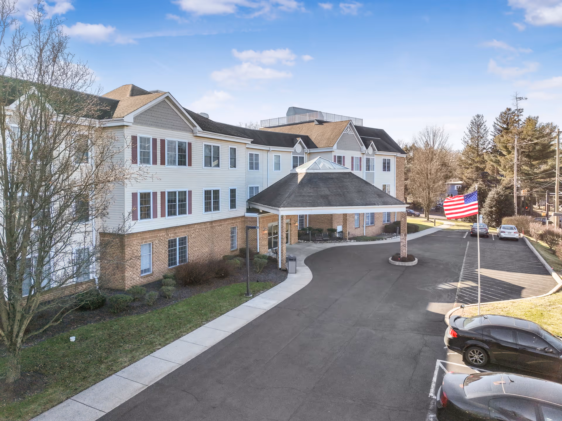 Exterior view of a multi-story senior living facility building with a covered entrance driveway, several parked cars, an American flag on a flagpole, and trees surrounding the property under a partly cloudy sky.