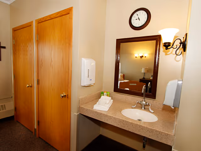 Interior view of a room corner featuring a beige countertop with a built-in sink, a wall-mounted soap dispenser, a mirror with a dark wooden frame, a wall clock above the mirror, and two wooden doors on the left side. A wall sconce light fixture is mounted next to the mirror, and folded white towels and small toiletry items are placed on the countertop.