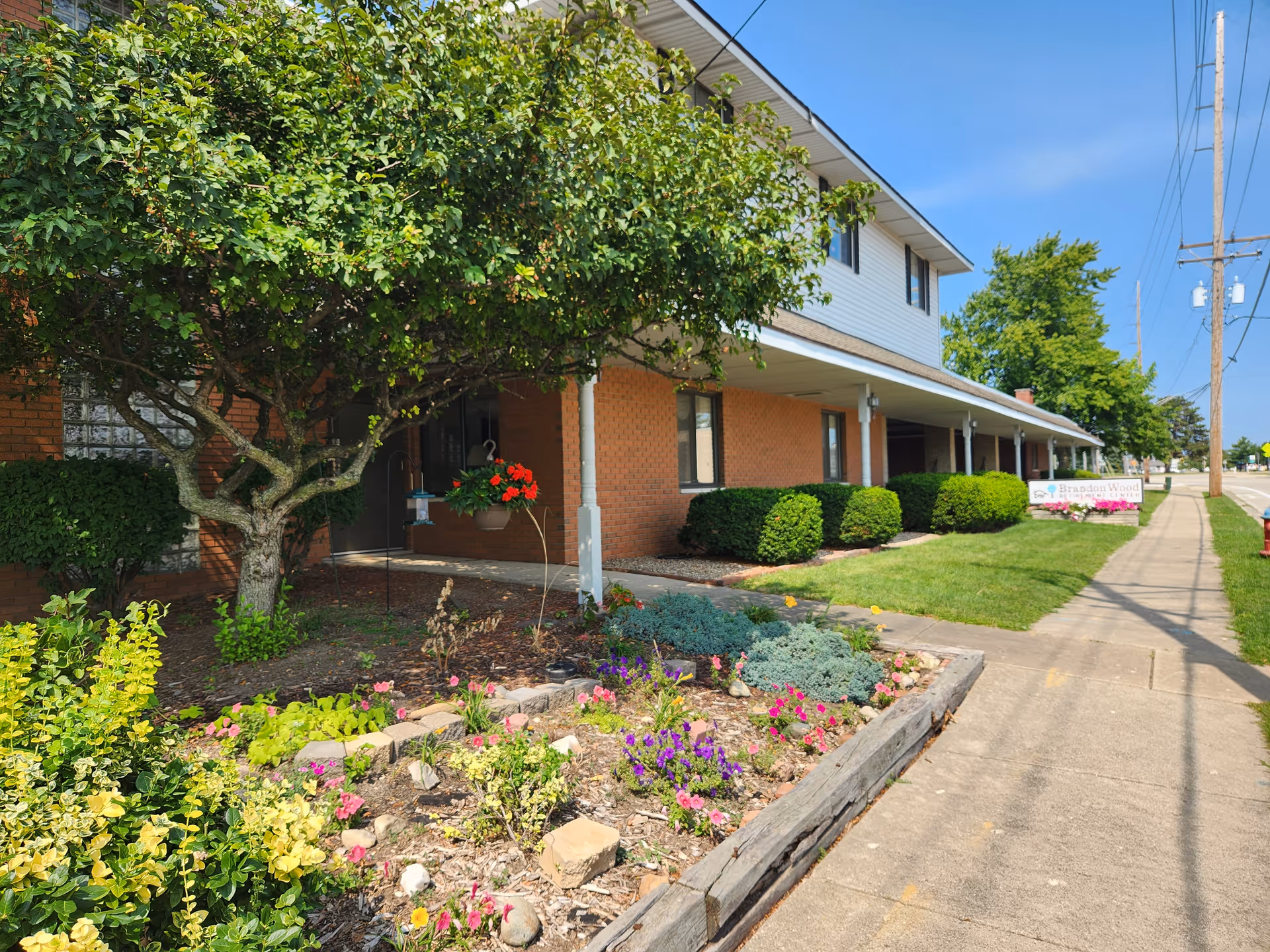 Exterior view of Brandon Wood Retirement Center showing a two-story brick and siding building with a covered walkway. There is a garden with various flowers and shrubs in the foreground, a tree providing shade near the entrance, and a sidewalk running alongside the building. The sky is clear and blue.