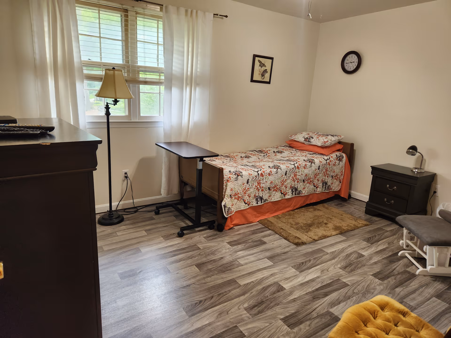 A sparsely furnished bedroom in an assisted living facility with a single bed, dresser, and window.