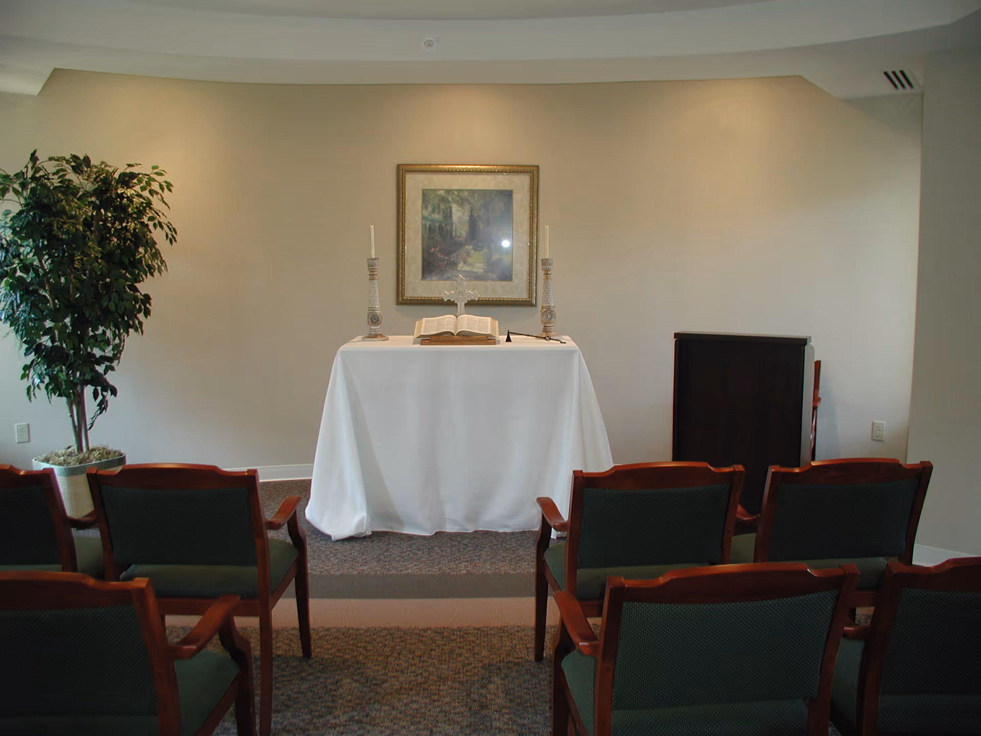 Small chapel or prayer room with rows of green cushioned wooden chairs facing a white-covered altar table. On the altar are two tall candlesticks, an open Bible, and a decorative cross. A framed picture hangs on the beige wall behind the altar, and a potted plant is placed to the left side of the room.