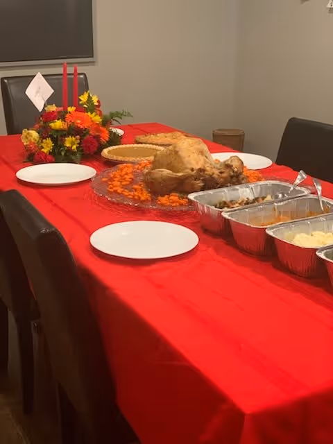 A dining table covered with a red tablecloth set with a roasted turkey on a platter surrounded by sliced carrots, several aluminum trays with side dishes, empty white plates, and a floral centerpiece with red candles in a simple dining room.