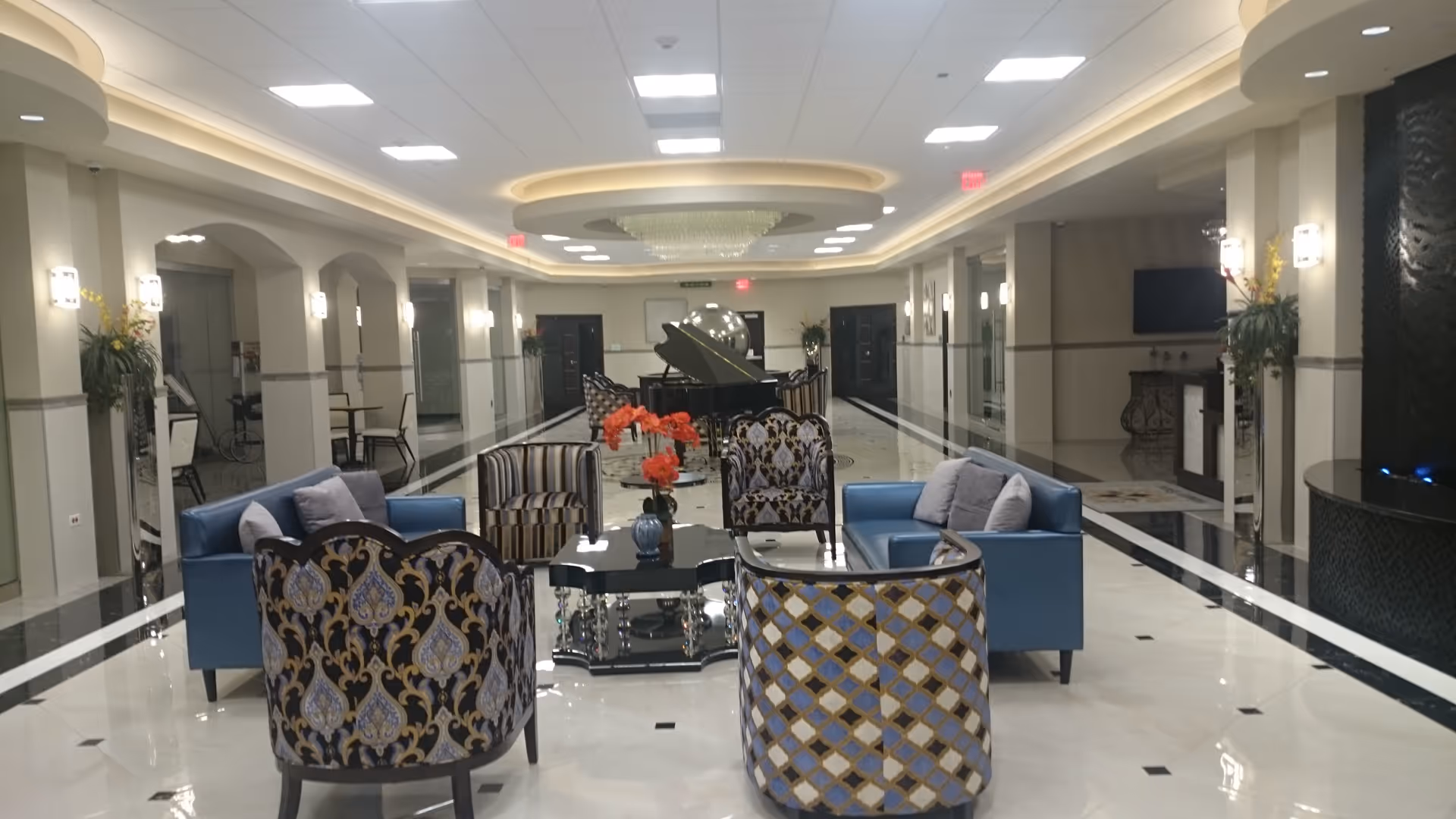 Luxurious hotel-style lobby with blue chairs, patterned seating, a central coffee table, and a grand piano.