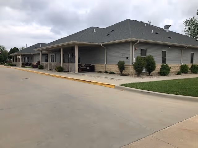 Exterior view of a single-story building with gray siding and a dark gray roof, featuring a covered porch area with seating. The building is surrounded by a concrete driveway and landscaped with small bushes and grass under a cloudy sky.