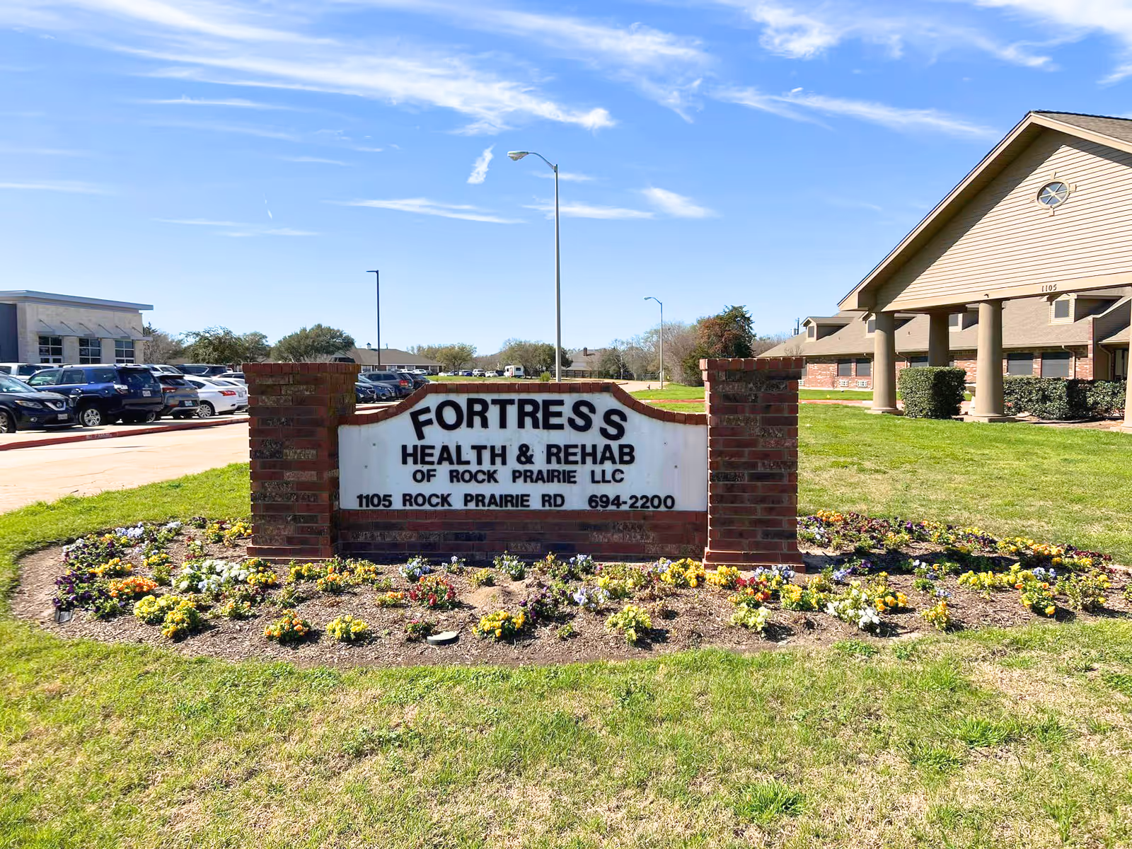 Front exterior of Fortress Nursing and Rehabilitation facility with brick sign and landscaped flower beds.