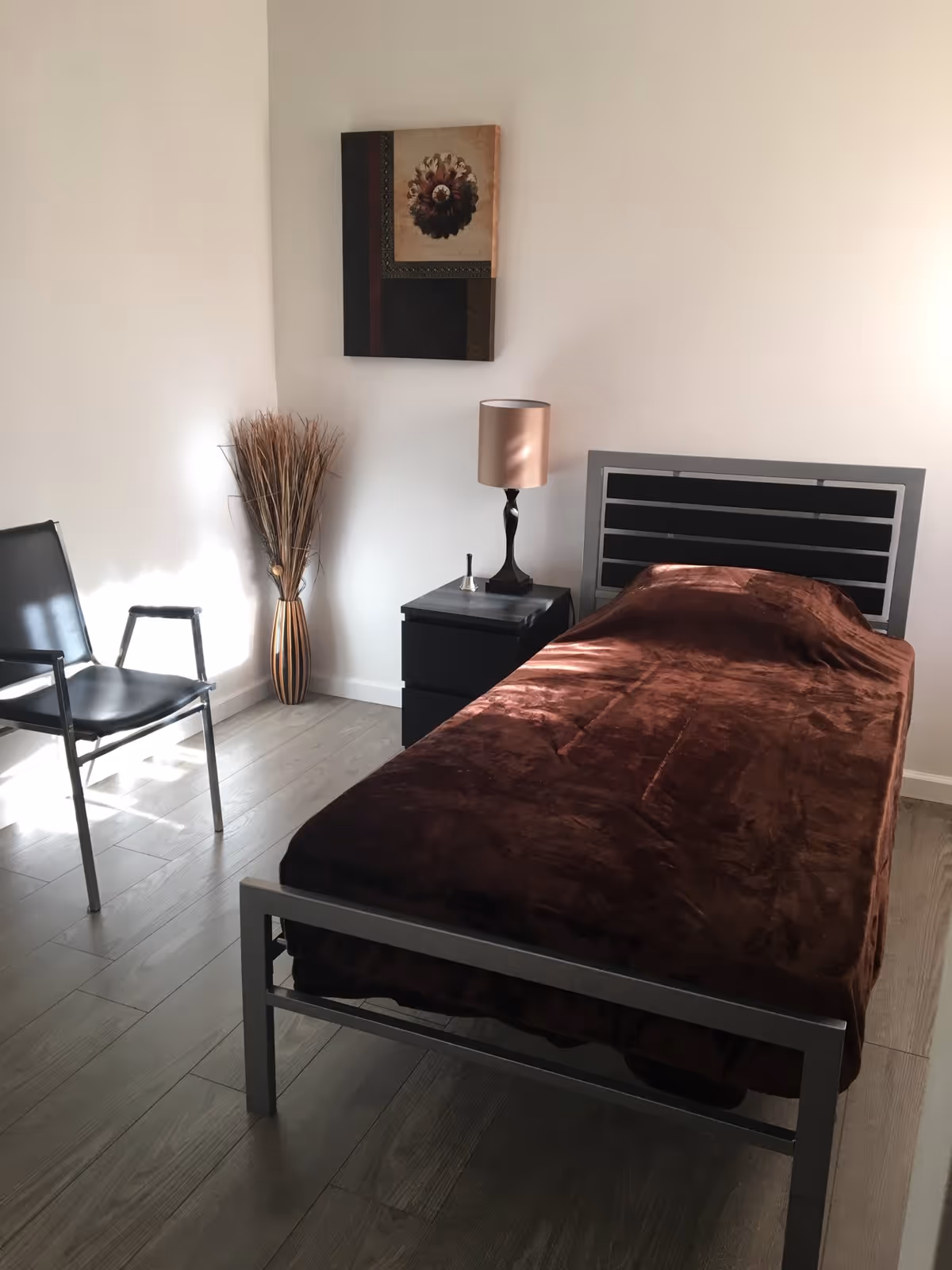 A simple bedroom with a single metal-framed bed covered with a brown blanket, a black nightstand with a lamp and a small bell on it, a black chair, a tall vase with dried decorative grass, and a framed artwork hanging on a white wall.