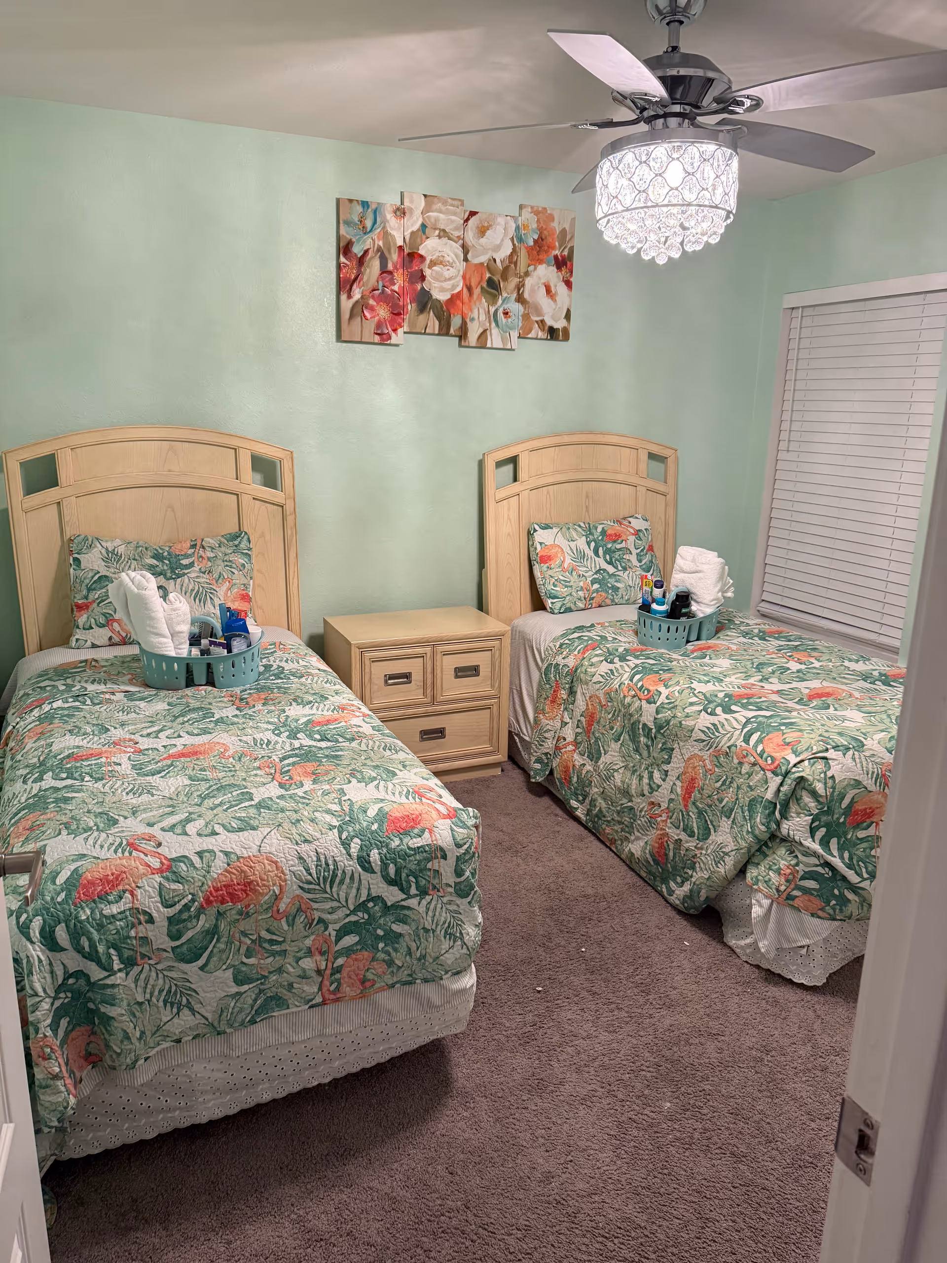 Two twin beds with tropical flamingo quilts, a shared nightstand, mint green walls, and ceiling fan.