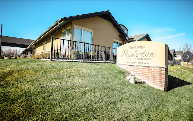 Exterior view of The Lodge At Riverton with a brick sign, railing, and manicured lawn.