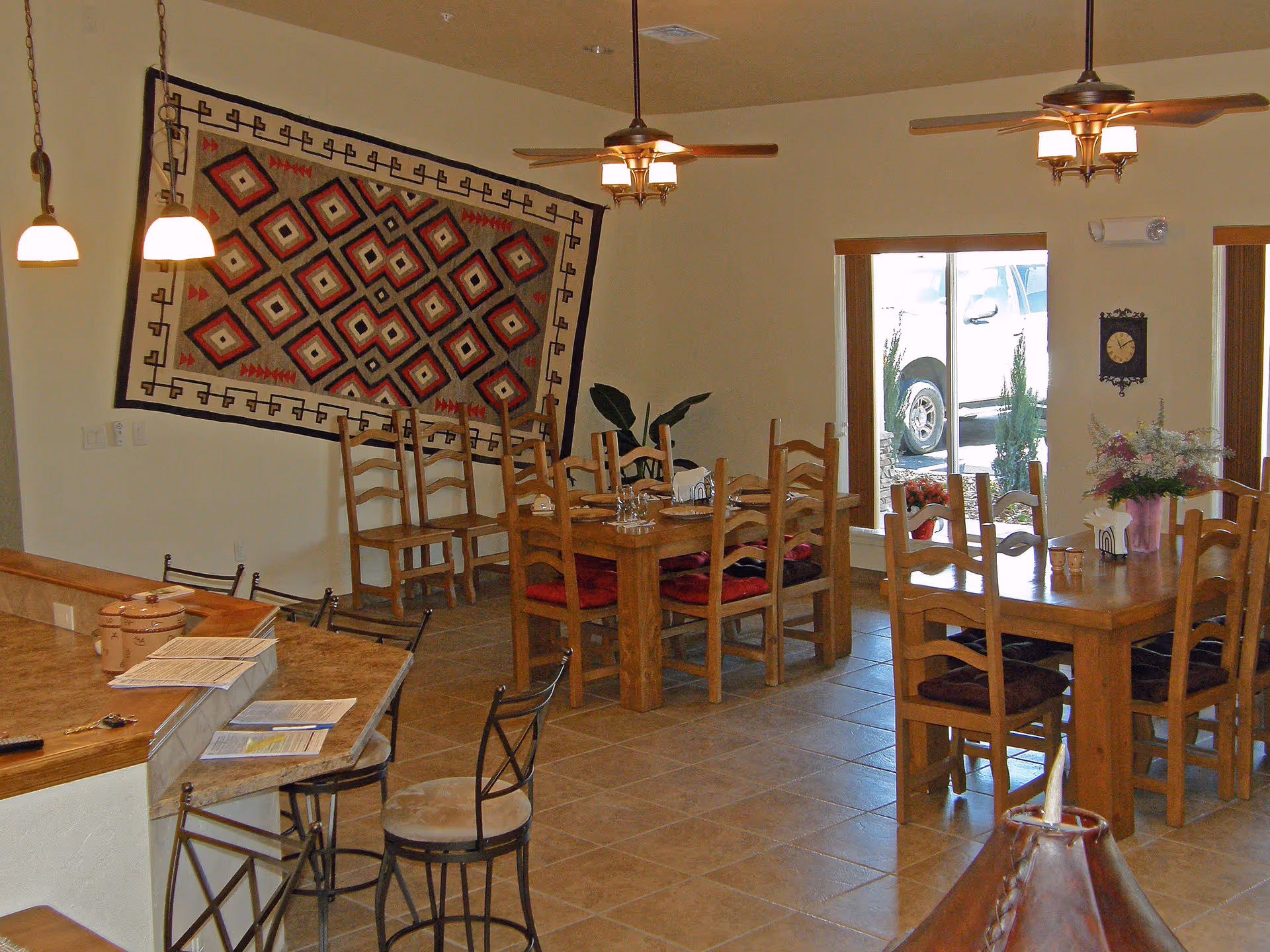 Interior view of a dining area with wooden tables and chairs, a decorative patterned wall hanging, ceiling fans with lights, and large windows showing a parked vehicle outside. There is a counter with bar stools and some papers on it.