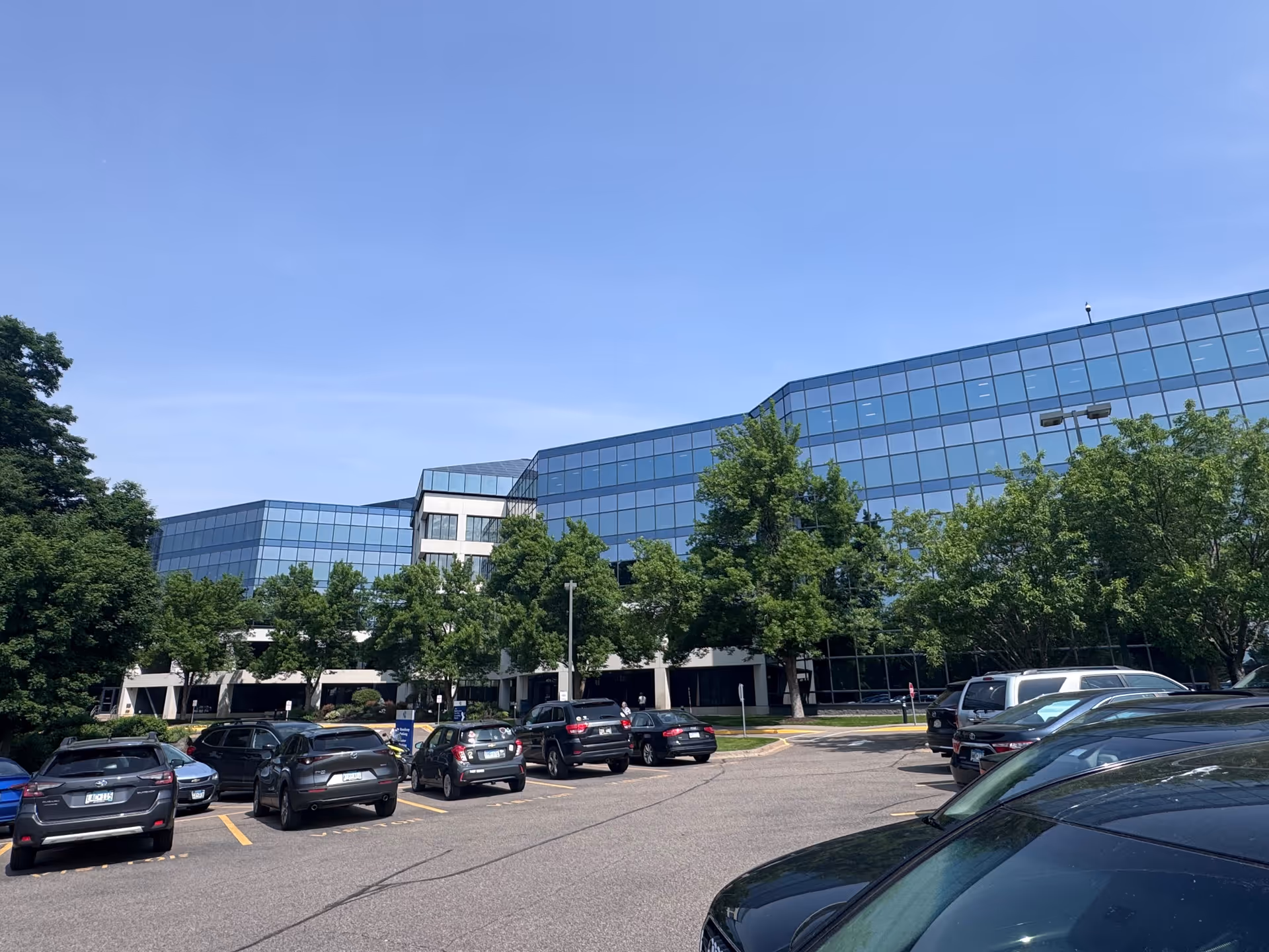Exterior view of a modern glass building with a parking lot and trees in front.