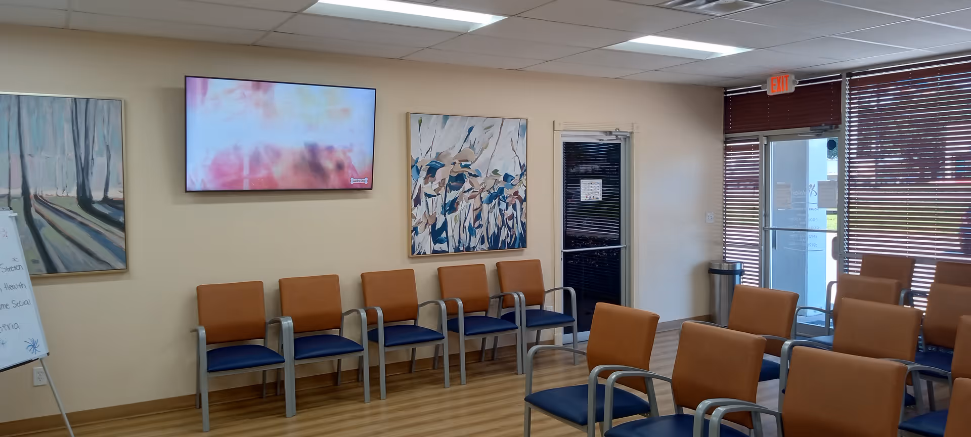 A waiting area with orange and blue chairs, wall art, a TV, and glass doors.