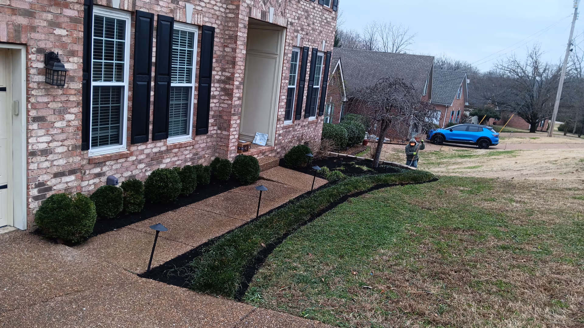 Brick residential building exterior with black shutters, a walkway, landscaped beds, and a distant worker.