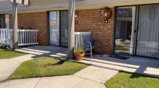 Brick-faced assisted living ground-floor units with sliding glass doors, small patios, chairs, potted plants, and a concrete walkway with lawns.