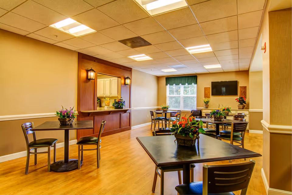 A well-lit dining room with several square tables and chairs arranged neatly. Each table has a floral centerpiece. The room features wood flooring, beige walls with white trim, a window with a green valance, and a wall-mounted flat-screen TV. There is a serving window with wooden trim and two wall-mounted lantern-style lights.