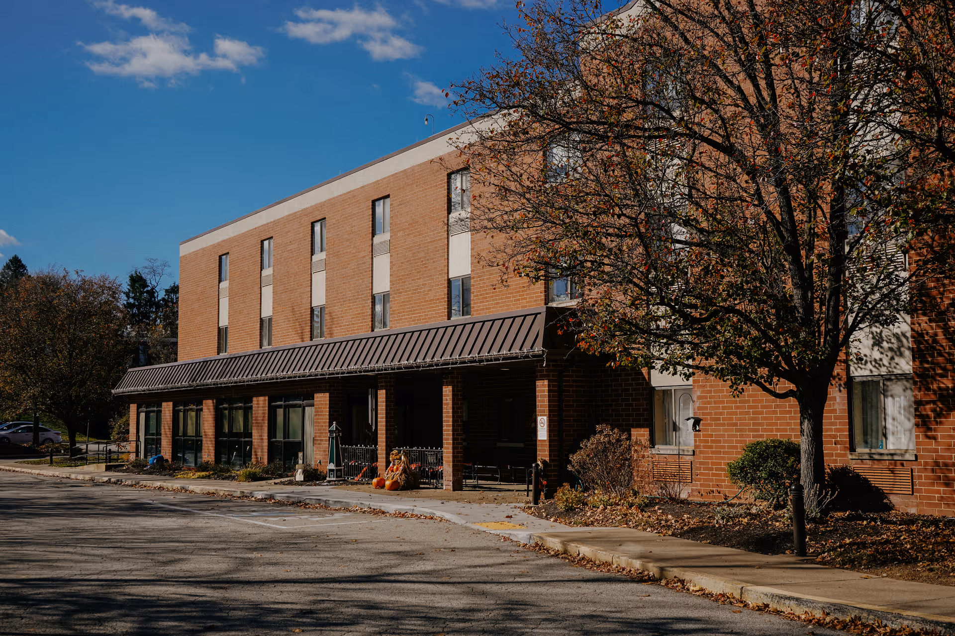 Exterior view of a three-story brick building with multiple windows, a covered entrance with a metal awning, and a tree with autumn leaves in front. The sky is clear and blue.