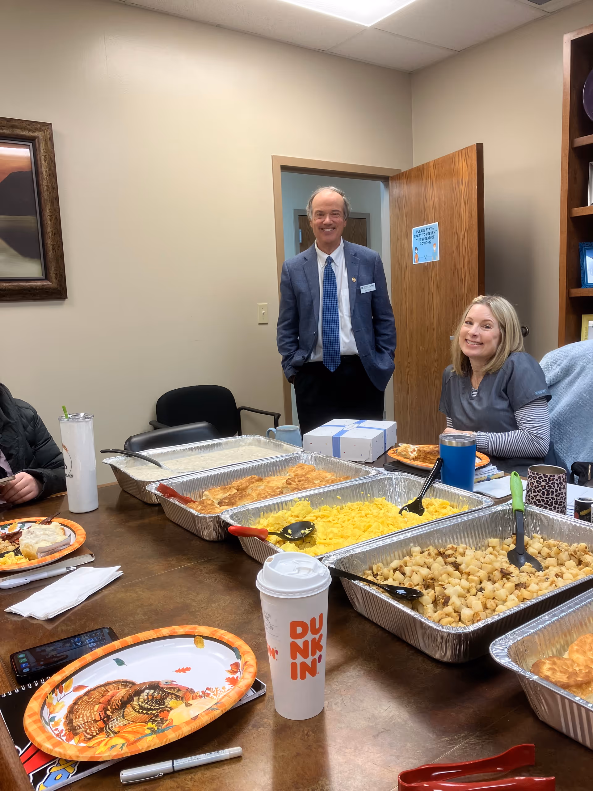 Man in suit and woman in scrubs near trays of food at a dining table.